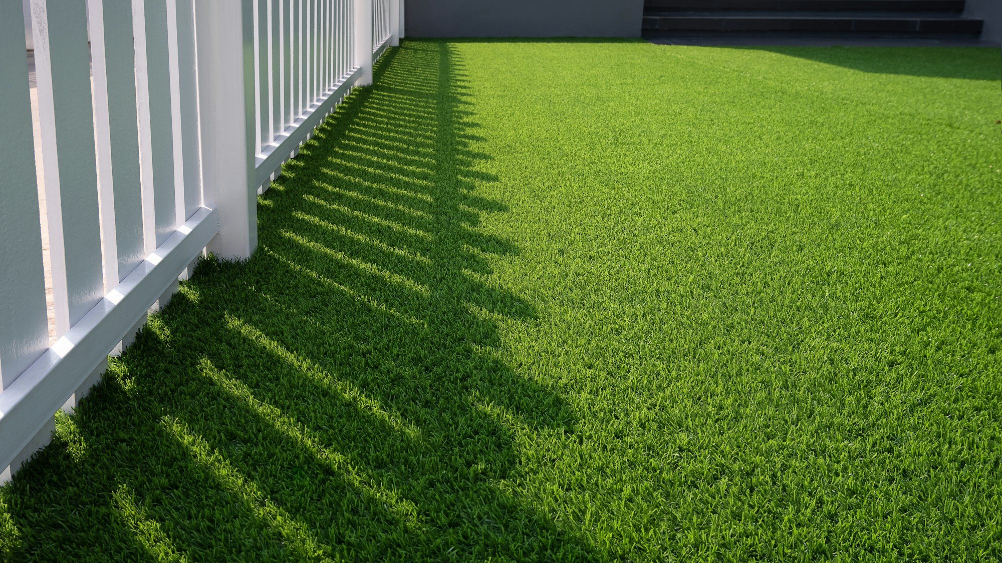Sunlight and shadow of white wooden fence on green artificial turf surface in front yard of home